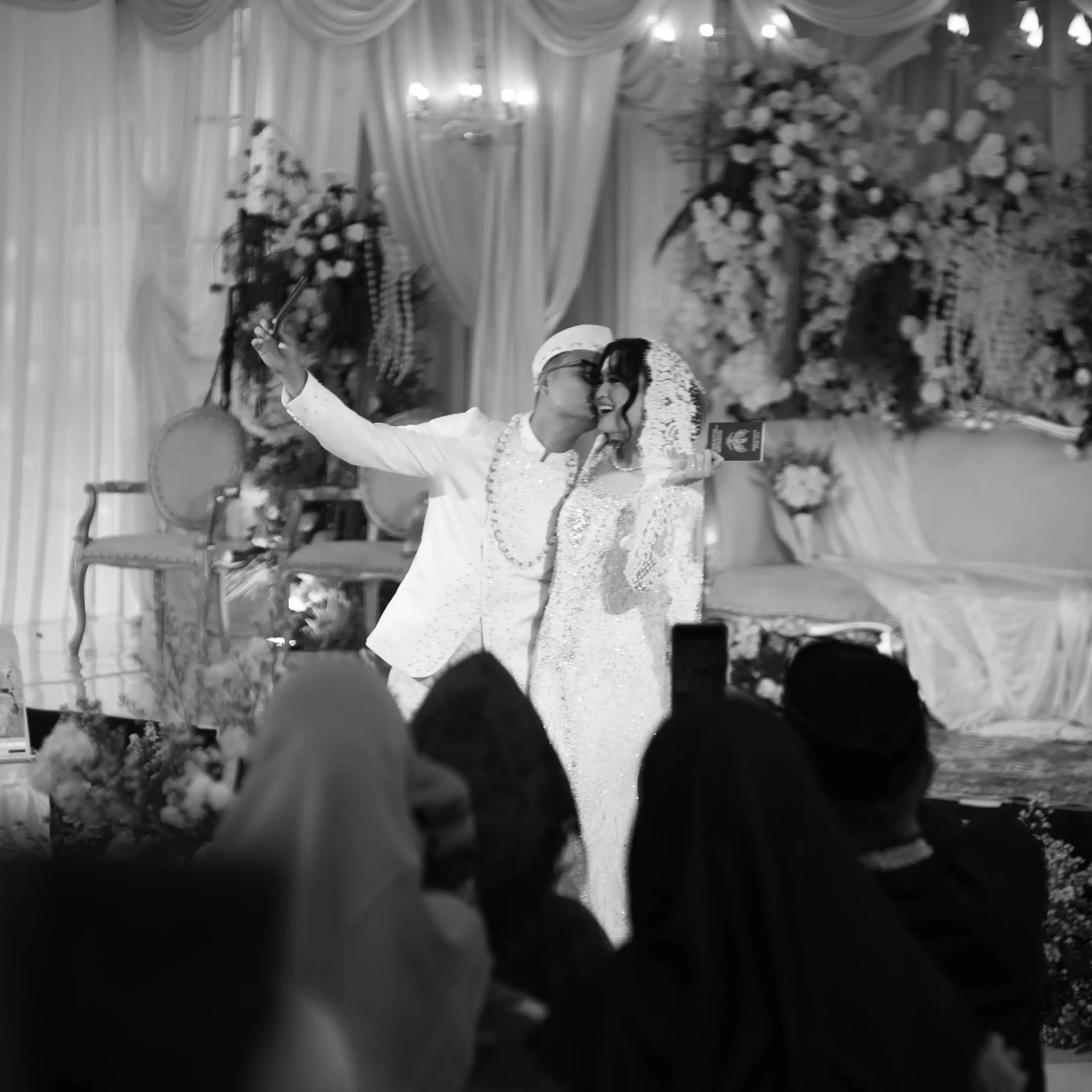 Foto hitam putih pengantin berswafoto di pelaminan berhias bunga