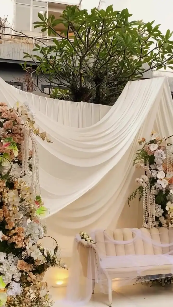Pelaminan dengan drape kain krem dan tiang bunga di taman terbuka