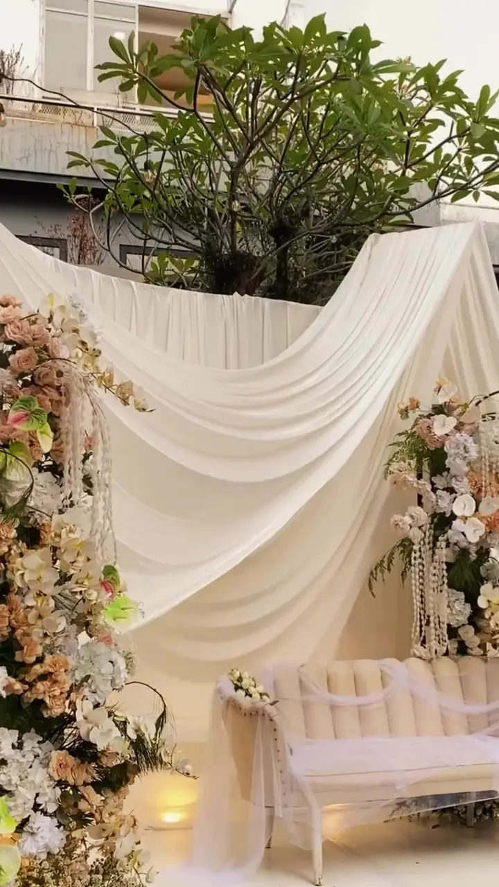 Pelaminan dengan drape kain krem dan tiang bunga di taman terbuka