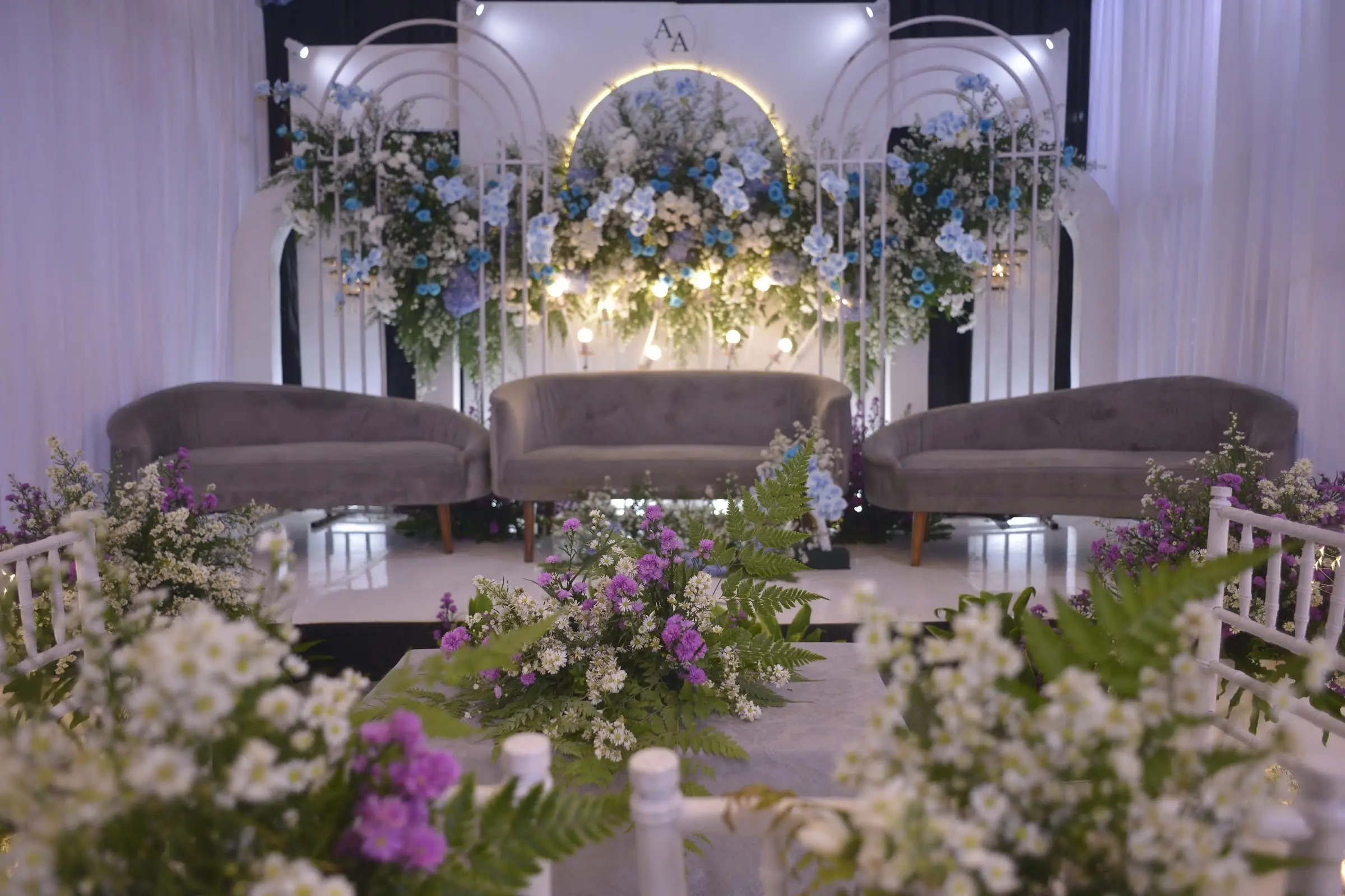 Pelaminan dengan tiga sofa abu-abu, backdrop lengkung putih, dan rangkaian bunga biru serta ungu