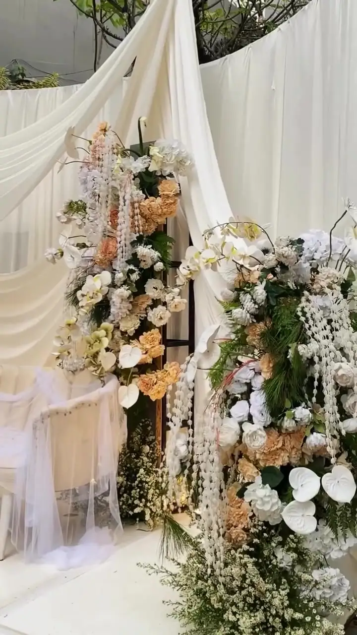 Rangkaian bunga putih, peach, dan anggrek menjuntai di sisi pelaminan berdrape kain