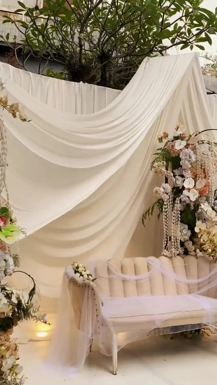 Sofa pelaminan putih di bawah drape kain krem dengan rangkaian bunga peach dan lampu lantai