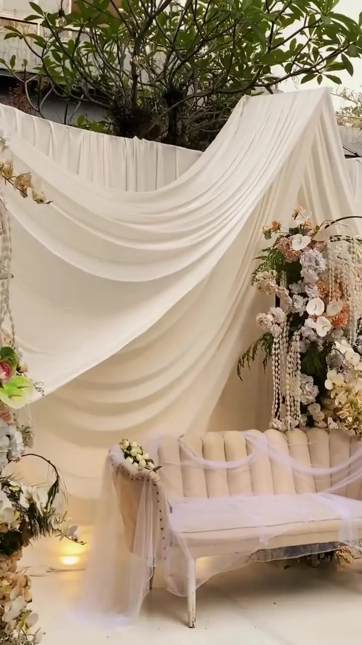 Sofa pelaminan putih di bawah drape kain krem dengan rangkaian bunga peach dan lampu lantai