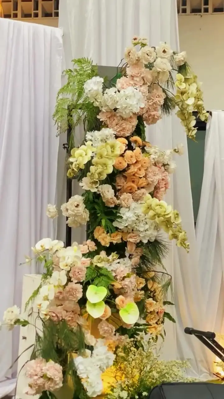 Tiang bunga tinggi berisi mawar peach, hortensia, dan anthurium hijau di depan kain putih